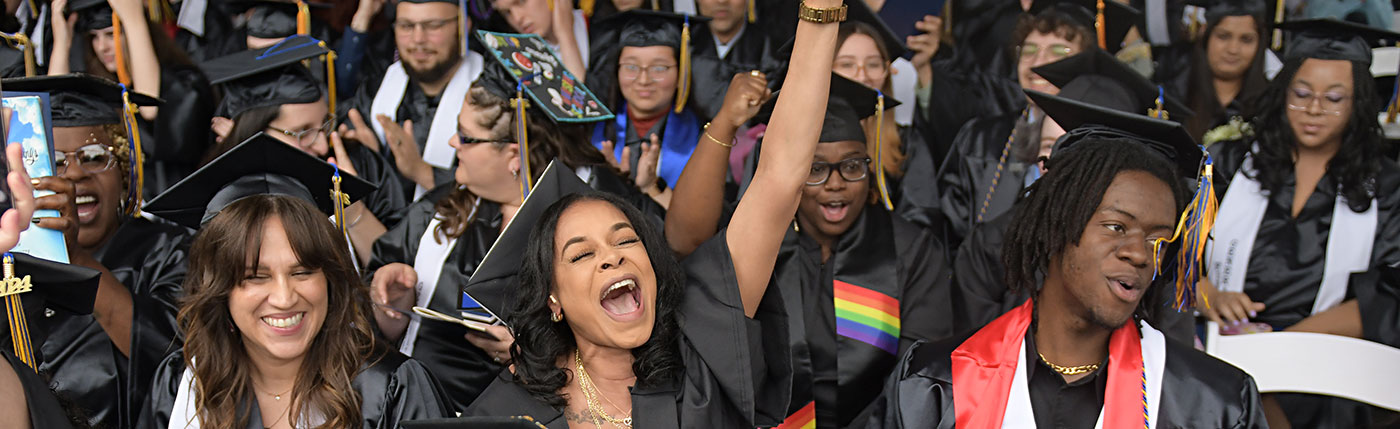 SUNY Schenectady graduates in their caps and gowns, one grad in the foregroud holding her diploma aloft with a look of joy on her face.