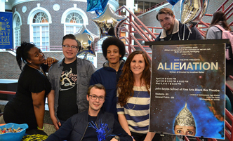 Students gather around a table and a large poster for an upcoming theater performance.