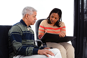 A community health worker(CHW) sitting with an older man, the CHW is writing in a notebook.