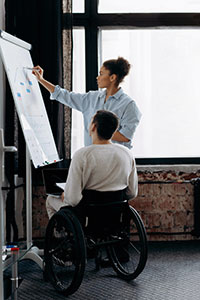 Two people working at an easel, one in a wheelchair with a laptop, the other standing writing on the paper. 