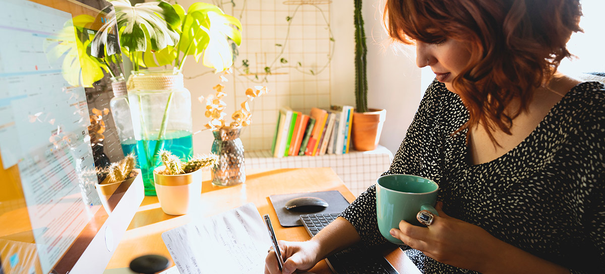 Woman, holding a coffee cup, sitting at a desk in a home, computer and notebook in front of her.